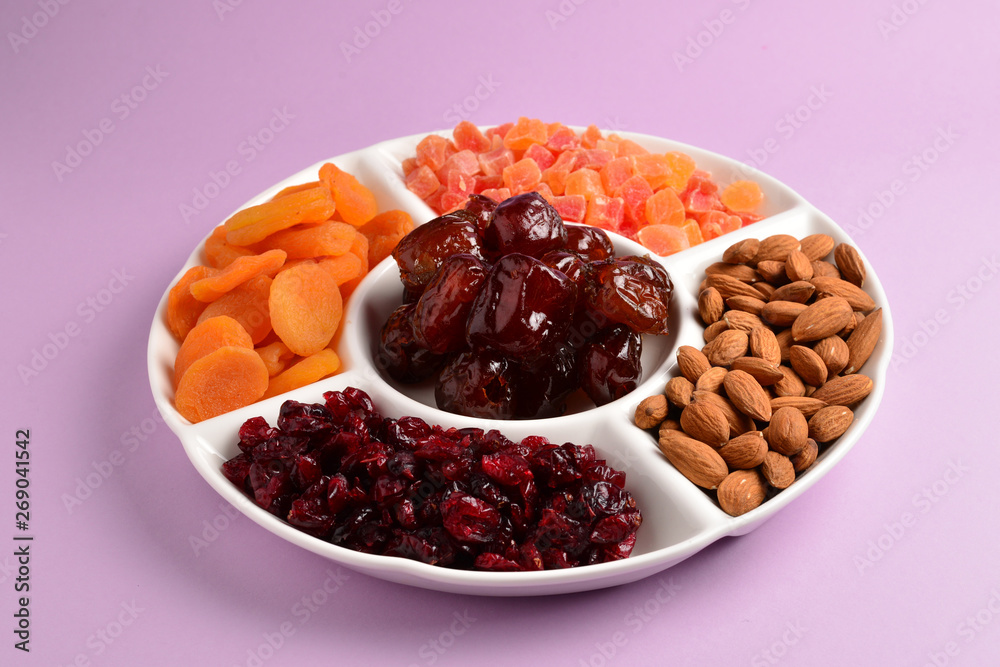 Mix of dried fruits and nuts on a white plate. Apricot, almond, raisin, dates fruit. On a lilac background. Space for text or design. Mix of dried fruits and nuts on a white plate. Apricot, almond, raisin, dates fruit. On a lilac background. Space for text or design.
