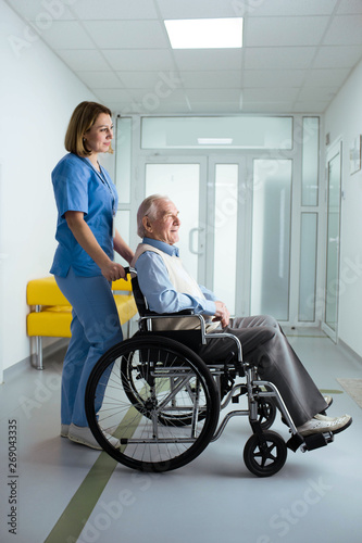 friendly nurse pushing senior patient in wheelchair at hospital corridor