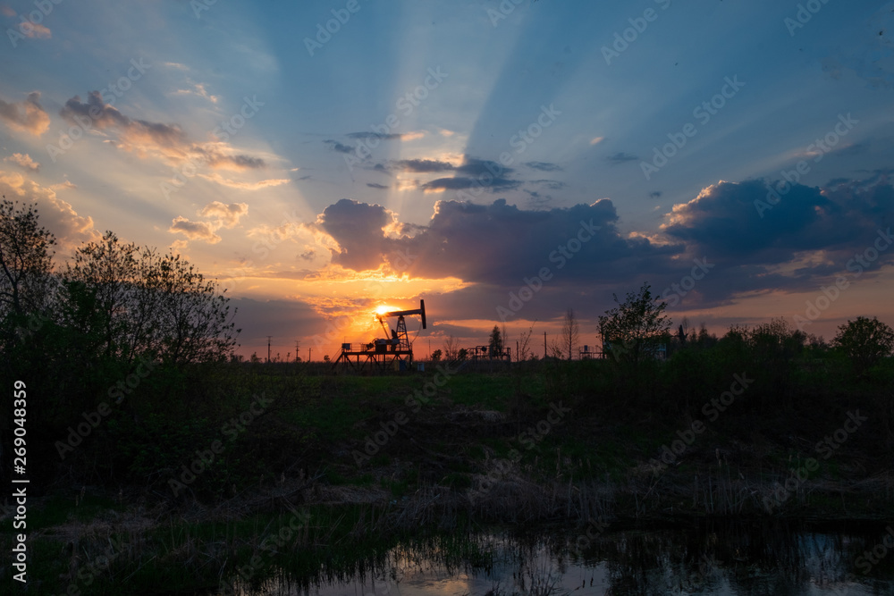 Fototapeta premium Equipment for oil and gas production works at sunset in the rays and glare of the sun next to the river. Oil well at sunset. Oil production with rocker at sunset. Rocking oil