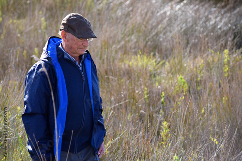 Hombre mayor con gorra paseando por el campo y observando el paisaje en un día soleado.