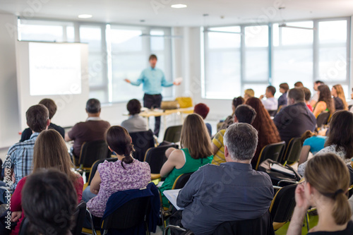 Business and entrepreneurship symposium. Speaker giving a talk at business meeting. Audience in conference hall. Rear view of unrecognized participant in audience. photo