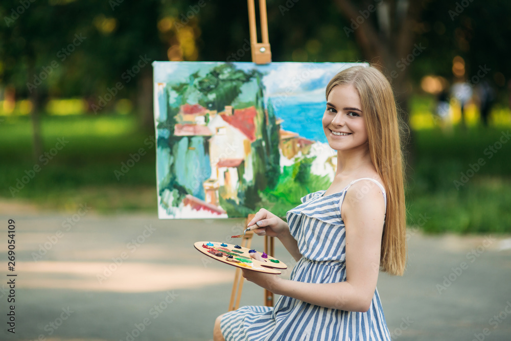Beautiful blond hair girl sitting on stool and draws a picture in the park using a palette with paints and a spatula