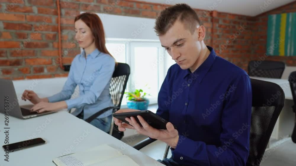 young caucasian coworkers sitting at the workplace in loft office. man using digital tablet reading documents employee with red hair typing on laptop young caucasian coworkers sitting at the workplace in loft office. man using digital tablet reading documents employee with red hair typing on laptop
