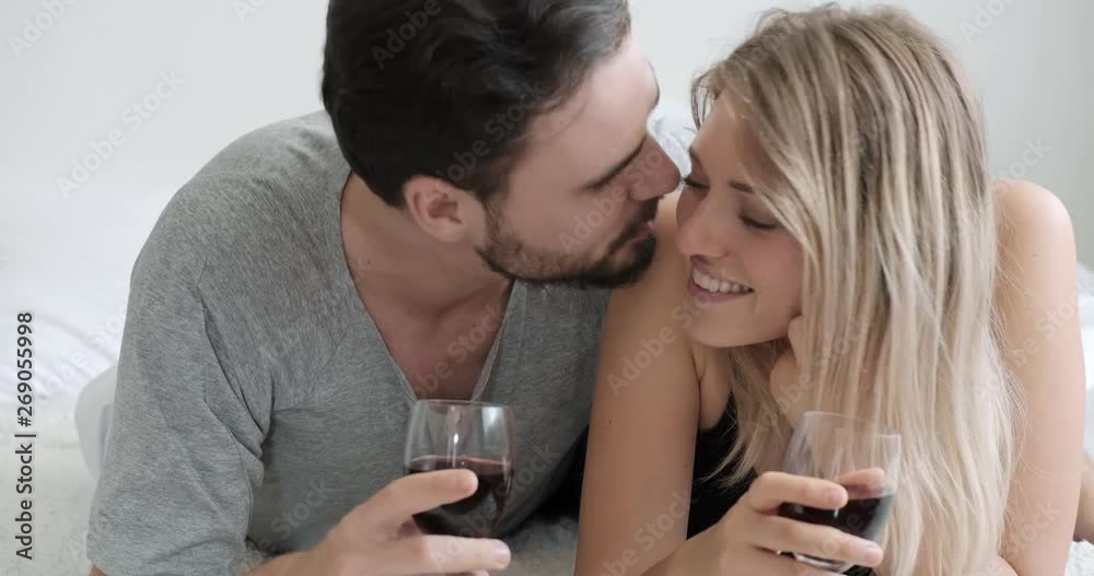 Young couple holding wine in bed and man kiss woman in the cheek. Caucasian man and woman. Romantic, anniversary, valentine concept. Young couple holding wine in bed and man kiss woman in the cheek. Caucasian man and woman. Romantic, anniversary, valentine concept.