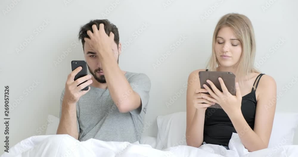 Couple in bed using mobile phone and tablet together, focus mood. Caucasian man and woman. Couple in bed using mobile phone and tablet together, focus mood. Caucasian man and woman.