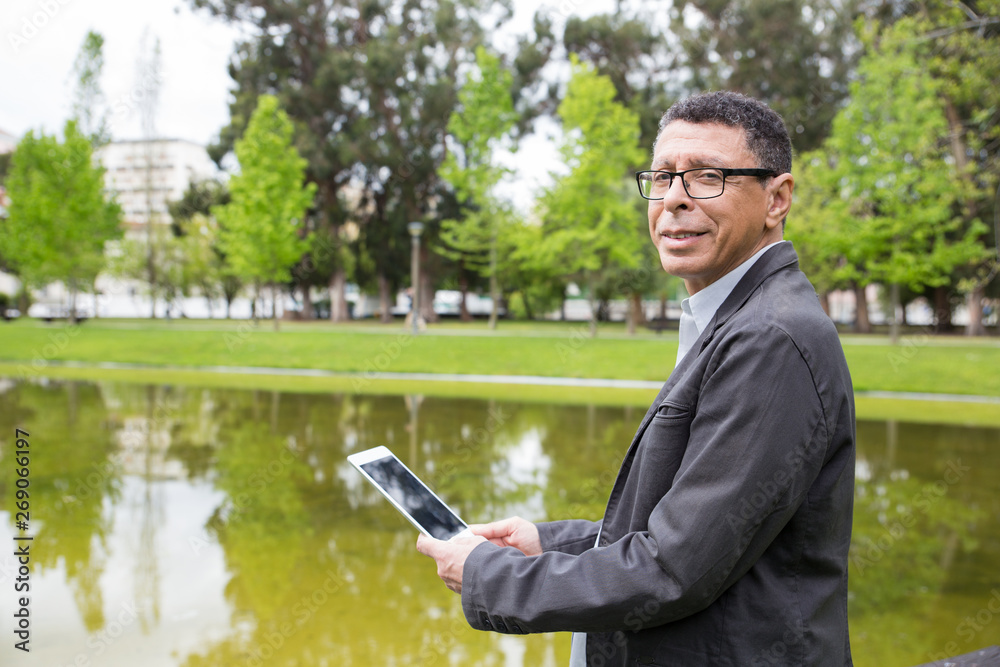 Smiling man using tablet and standing in city park. Guy wearing casual clothes and holding gadget with pond and green trees in background. Communication and nature concept. Side view. Smiling man using tablet and standing in city park. Guy wearing casual clothes and holding gadget with pond and green trees in background. Communication and nature concept. Side view.
