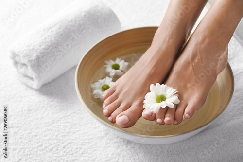 Closeup view of woman soaking her feet in dish with water and flowers on white towel, space for text. Spa treatment