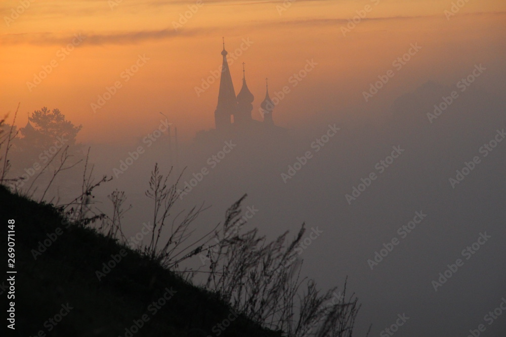 Silhouettes of church, trees, hill grass in misty morning 