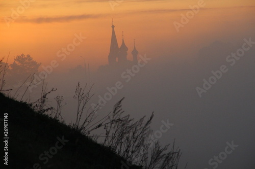 Silhouettes of church, trees, hill grass in misty morning  © Natalya
