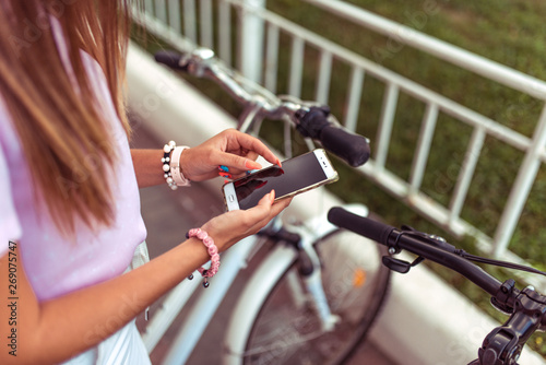 Girl in the summer in the city in hand phone, choice of route, background of the parking bicycles. Online application for renting bicycles. Female hands smartphone manicure bracelets and long hair.