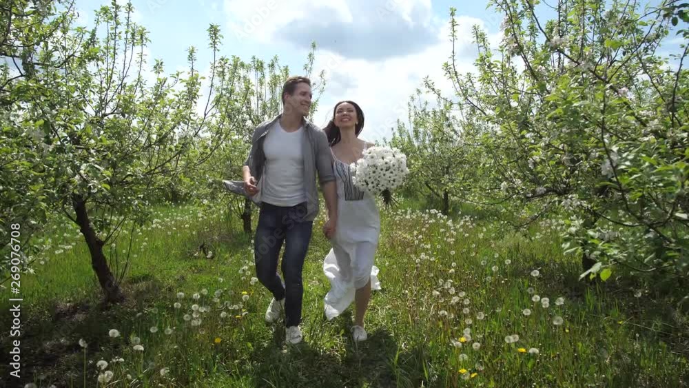 Joyful young mixed race couple in love running hand in hand in blooming apple orchard. Attractive groom and bride having fun running holding hands during romantic walk among flowering trees Joyful young mixed race couple in love running hand in hand in blooming apple orchard. Attractive groom and bride having fun running holding hands during romantic walk among flowering trees