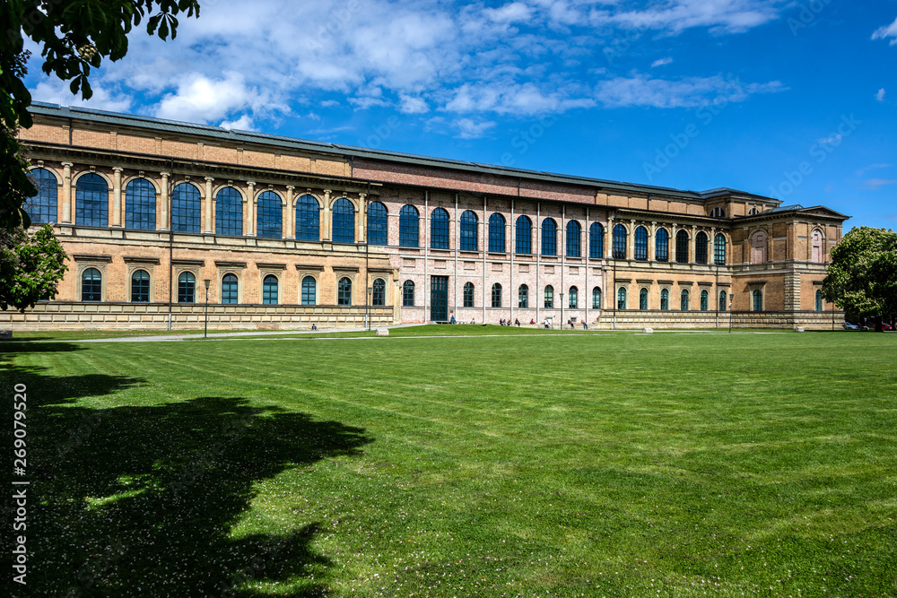 Germany, Munich: Famous Alte Pinakothek in the city center of the Bavarian capital seen from Gabelsbergerstrasse with people, public park, green grass and blue cloudy sky in background.