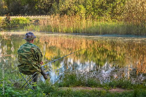 fisherman dressed in camouflage and rubber boots sitting with a fishing rod on the shore of the lake in the woods