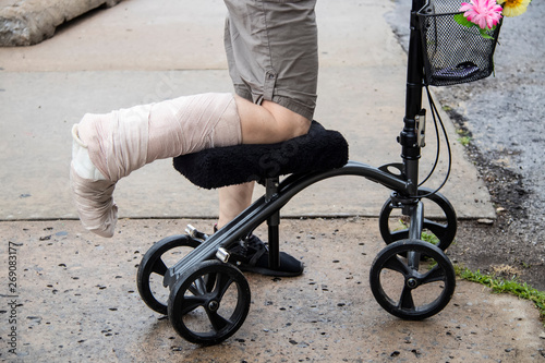 Woman with wrapped leg on steerable knee walker scooter with flowers on basket and mobile phone inside on rough broken cement surface - cropped photo