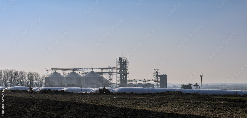 View of modern sugar factory (refinery) in the distance with industrial silos (large metal tanks for sugar or silage storage) and equipment, long polymeric bags for grain storage in the foreground