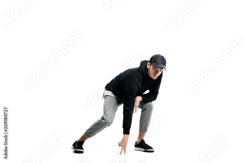 Young man wearing a black sweatshirt, gray pants, cap and sneakers dancing street dances on a white background
