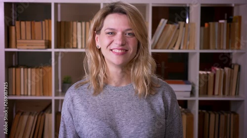 Cute middle-aged blonde teacher puts thumb-up sign to show likeness and respect into camera at the library.