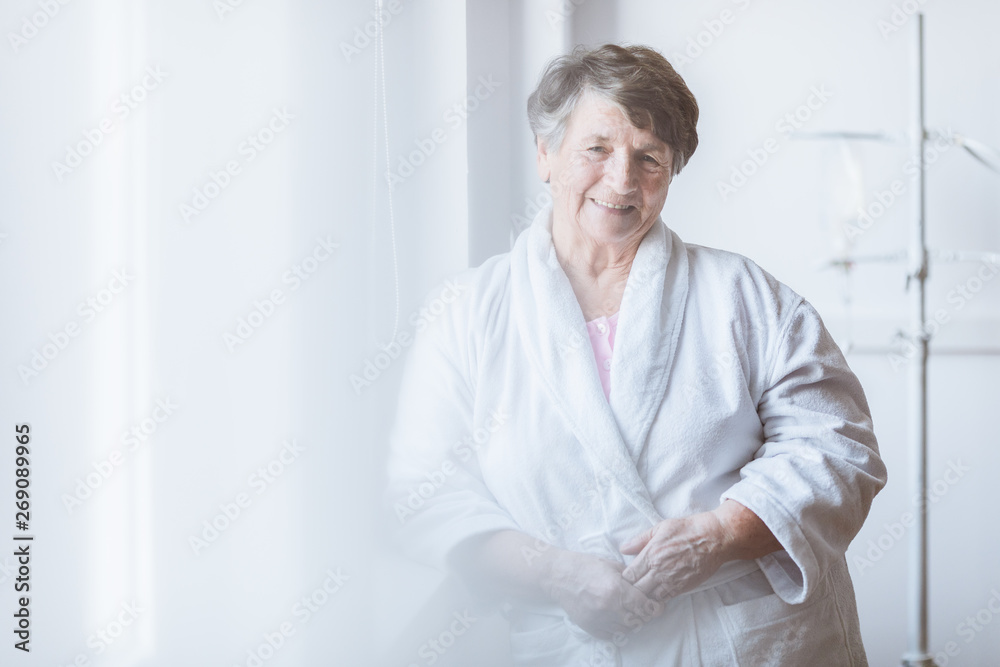 Senior grey lady wearing white bathrobe standing by the window at nursing home