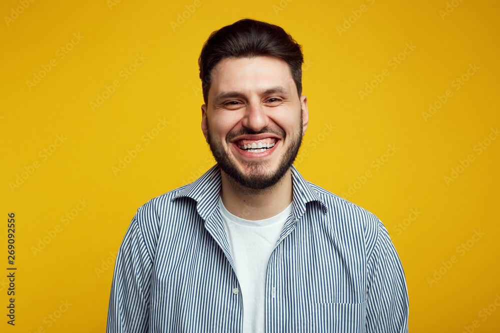Attractive bearded young man laughing out loud, smiling broadly, showing his white straight teeth against yellow background