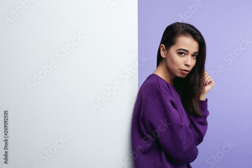 Pretty young girl, standing alongside empty white billboard wall isolated over purple background.