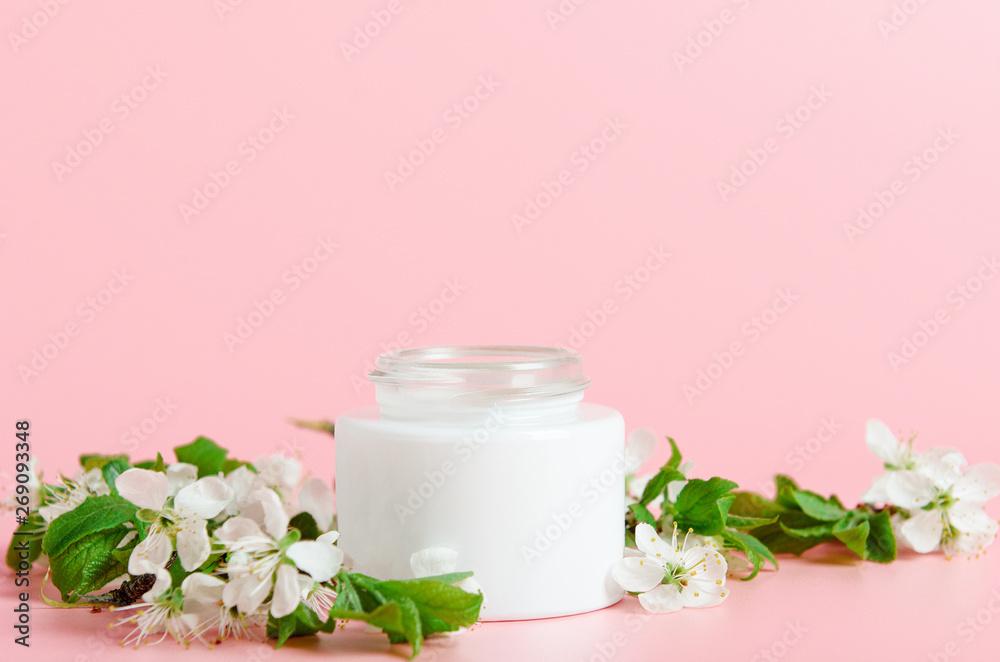 Face cream in white jar on a pink background with white flowers. Concept natural cosmetics, organic beauty. Copy space.