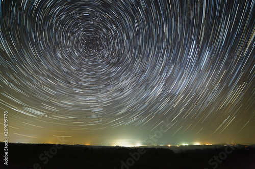 Trail of stars in the night sky around the North Star. Photographed with a long exposure in the background of the illuminated city.