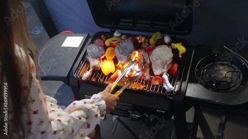 Sexy young girl prepares steaks and grilled vegetables on the roof of the house overlooking the night city, the wind lifts her dress and underwear can be seen