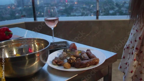 Sexy young girl prepares steaks and grilled vegetables on the roof of the house overlooking the night city, the wind lifts her dress and underwear can be seen