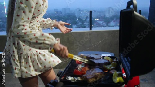Sexy young girl prepares steaks and grilled vegetables on the roof of the house overlooking the night city, the wind lifts her dress and underwear can be seen