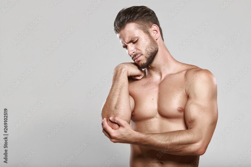 Fototapeta premium Muscular guy holding his injured elbow on gray background. Young strong man feeling the pain in his elbow after exercise. Healthcare, medical and sickness concept. Sports and muscle injury.