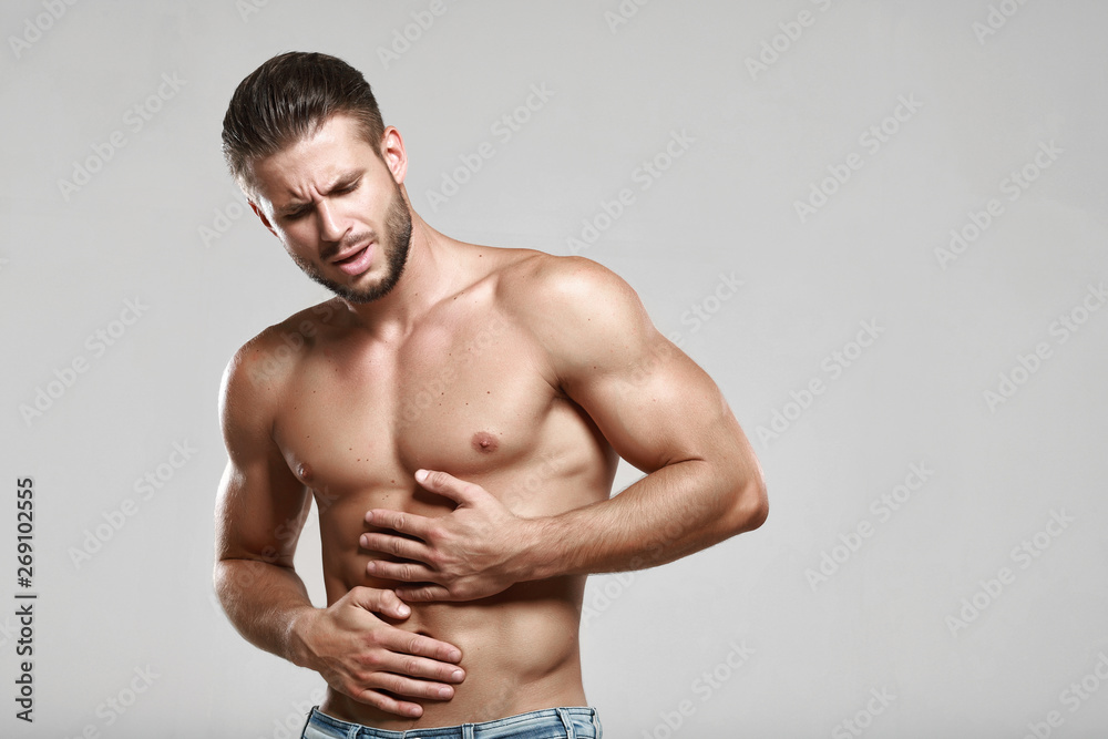 Young man suffering from stomach ache. Causes of abdominal pain include gastritis, stomach ulcer, food poisoning, diarrhea or IBS. Gray background. Healthcare, medical and sickness concept. Young man suffering from stomach ache. Causes of abdominal pain include gastritis, stomach ulcer, food poisoning, diarrhea or IBS. Gray background. Healthcare, medical and sickness concept.