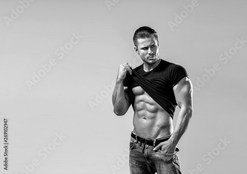 Muscular model sports young man in blue jeans raised his hand t-shirt and showing his press on a grey background. Black and white fashion portrait of sporty healthy strong muscle guy. Sexy torso.