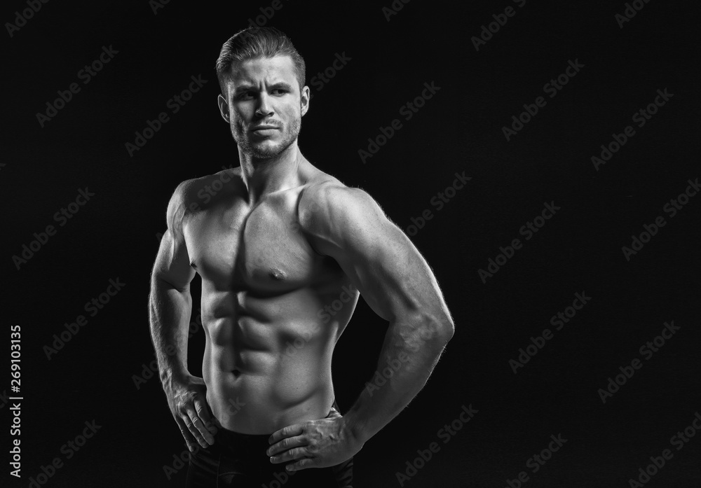 Muscular model young man on dark background. Black and white fashion portrait of strong brutal guy with modern trendy hairstyle. Sexy naked torso six pack abs. Male flexing his muscles. Sport concept.