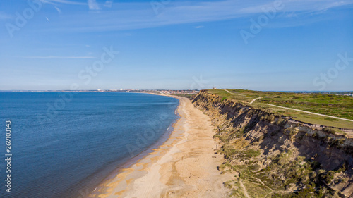 An aerial view of a beautiful beach with crystal blue water along a hill with cliff under a majestic blue sky and some white clouds