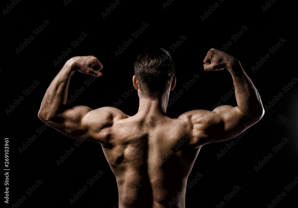 Muscular model sports young man on a dark background. Sporty healthy strong  muscle guy showing his back and big shoulders,biceps, triceps. Sexy male  flexing his muscles. Workout bodybuilding concept. Stock Photo