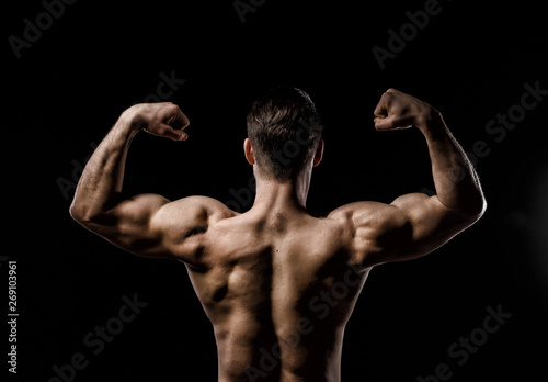 Muscular model sports young man on a dark background. Sporty healthy strong muscle guy showing his back and big shoulders,biceps, triceps. Sexy male flexing his muscles. Workout bodybuilding concept.