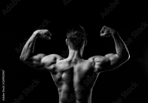 Muscular model sports young man on dark background. Sporty guy showing his back and big shoulders,biceps, triceps. Sexy male flexing his muscles. Workout bodybuilding concept. Black and white.
