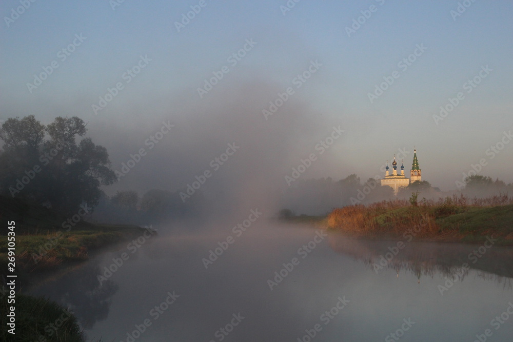 Fototapeta premium Misty cloud on river, green shore, trees, church in summer morning