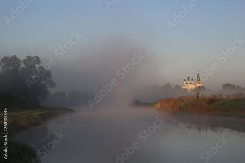 Misty cloud on river, green shore, trees, church in summer morning