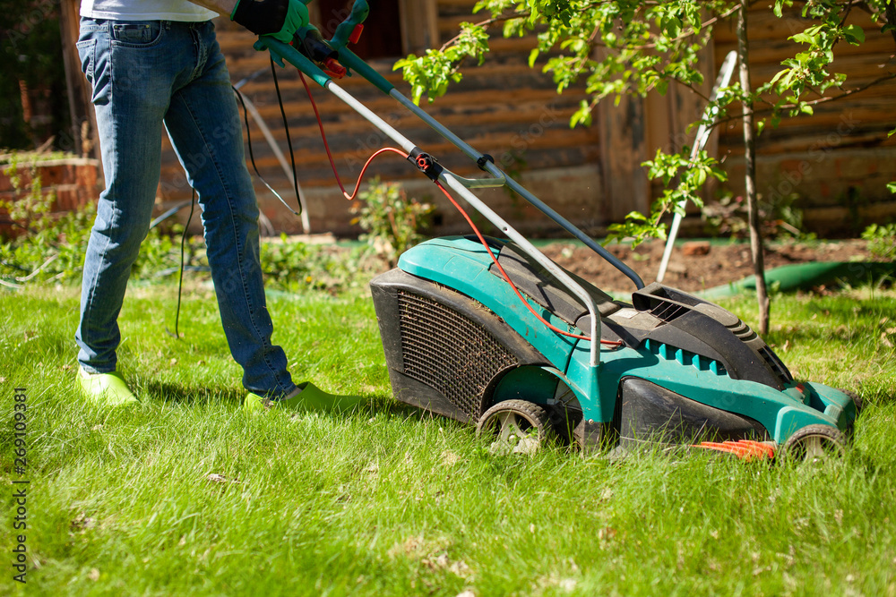 Fototapeta premium Young man mowing the lawn. Worker doing his job in backyard. Spending summer day in garden. Lawnmower standing on the background of private garden.