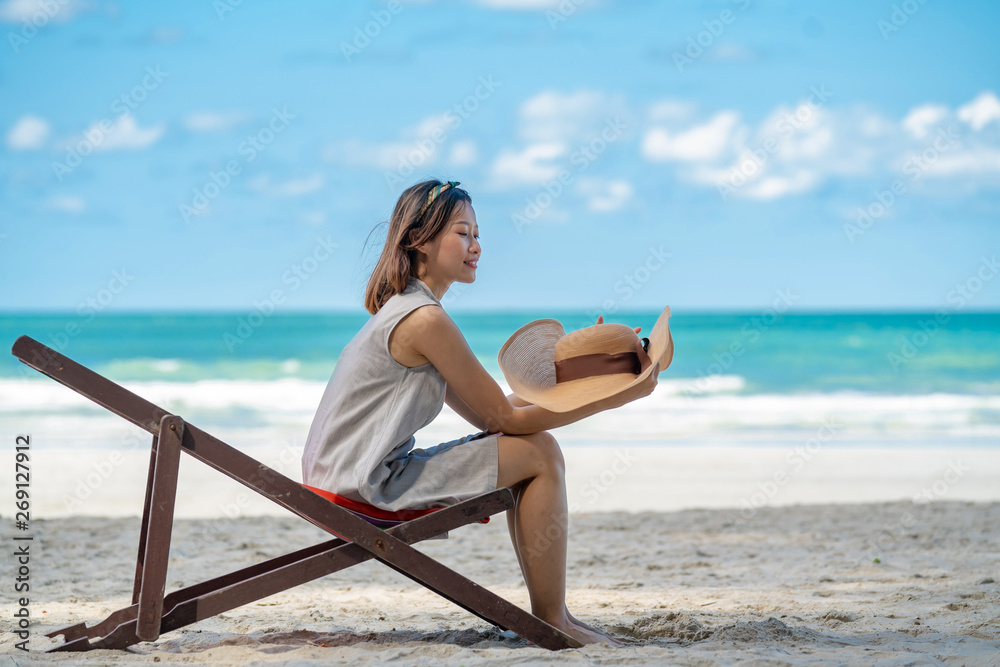 Selective focus of young asian adult traveler in casual wear sitting on beach chair on sand beach in tropical island in summer day holidays vacation with blurred blue sea, beach and sky backgrounds.
