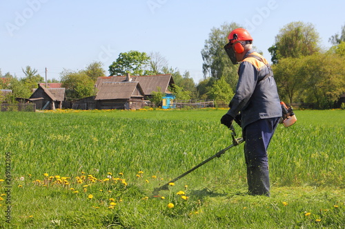 Russian farmer mowing the lawn with mower trimmer in a field on background of village houses on a Sunny summer day - working with garden tools