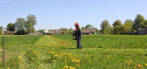 Farmer mows green grass with mower trimmer on the road in a field on the background of village houses and blue sky on a Sunny summer day - rural landscape