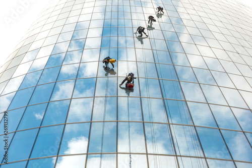 Five window washers work at a height on a high-rise building with a glazed facade against a blue sky with light clouds.