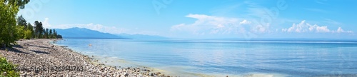 Panoramic view of Baikal Lake on a sunny summer day. Pebble beach and coastal grove. In the distance, tourists are resting. Beautiful water landscape