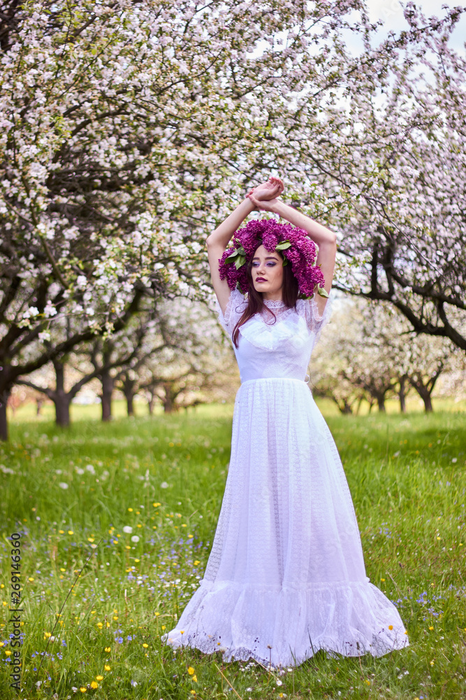 Young beautiful girl in a wreath of lilacs. White lace dress on the perfect figure of the model. Photo session in the spring apple orchard in the open.
