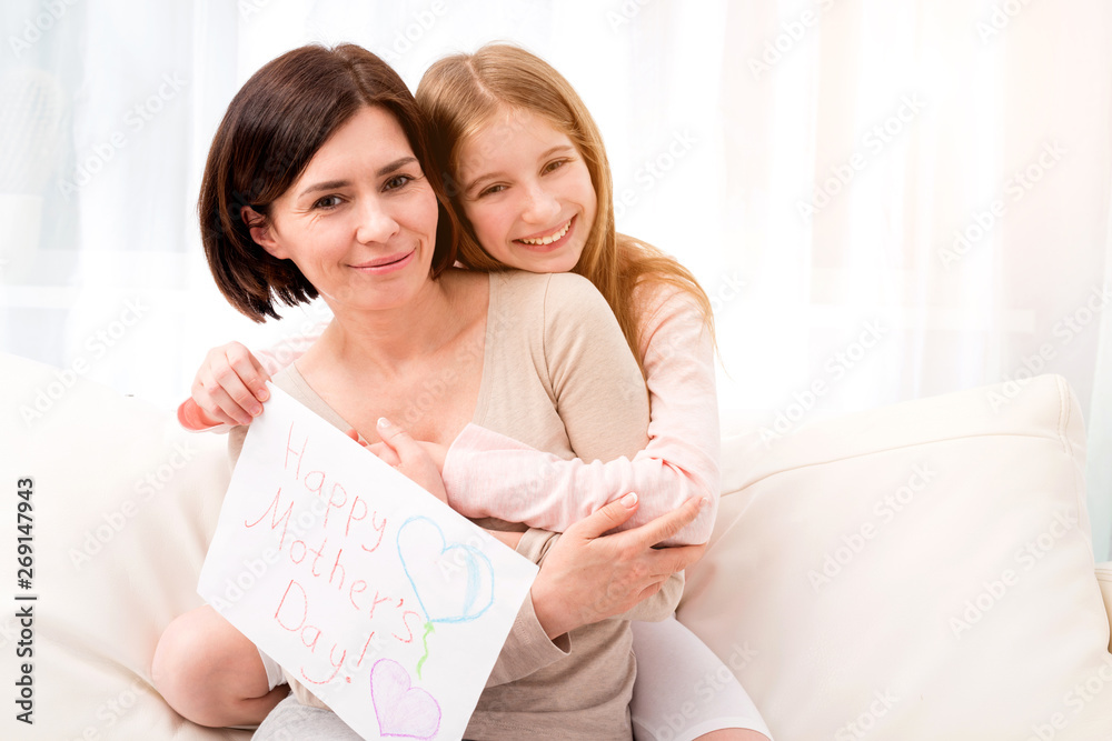 Lovely little girl hugging her mother and looking at camera. Girl holding creative handmade greeting card for mother. Daughter congratulates mom and gives her postcard for Happy Mother's day Lovely little girl hugging her mother and looking at camera. Girl holding creative handmade greeting card for mother. Daughter congratulates mom and gives her postcard for Happy Mother's day