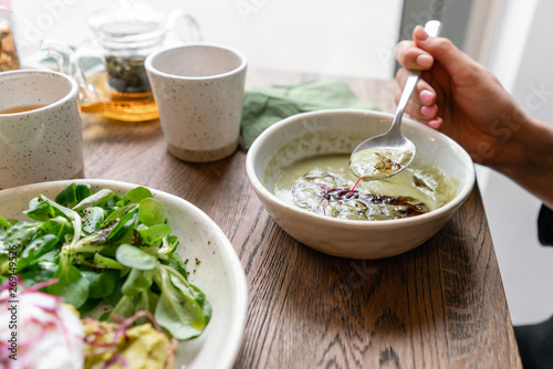 Woman eating fresh vegetable detox soup made of broccoli with with pumpkin seeds. In the foreground toast with avocado and poached egg, spinach salad.