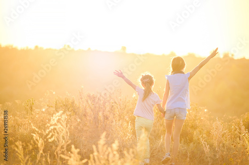 Little girls stand by holding hands looking on sunshine evening field with joyfully raised hands