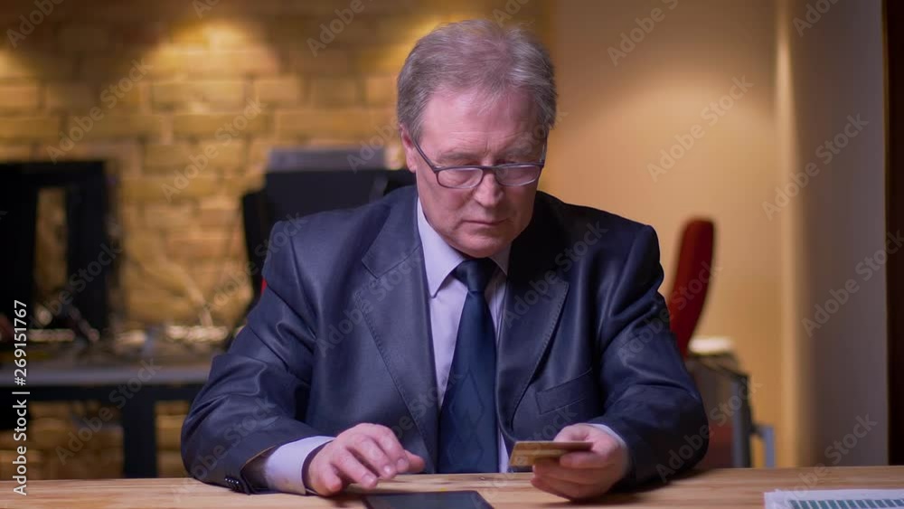 Portrait of senior businessman in formal costume making transactions with credit card and tablet in office. Portrait of senior businessman in formal costume making transactions with credit card and tablet in office.